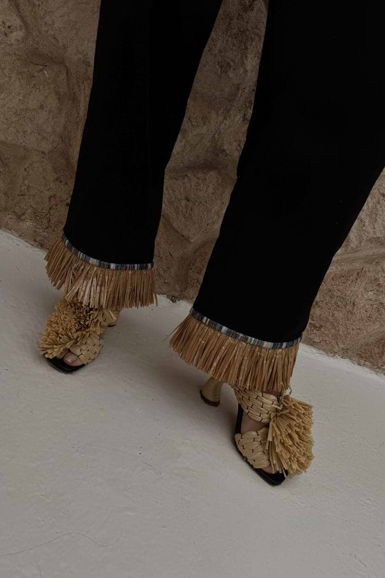 Black Raffia Marra Trousers featuring tan raffia fringe at the hem, styled with woven raffia sandals.