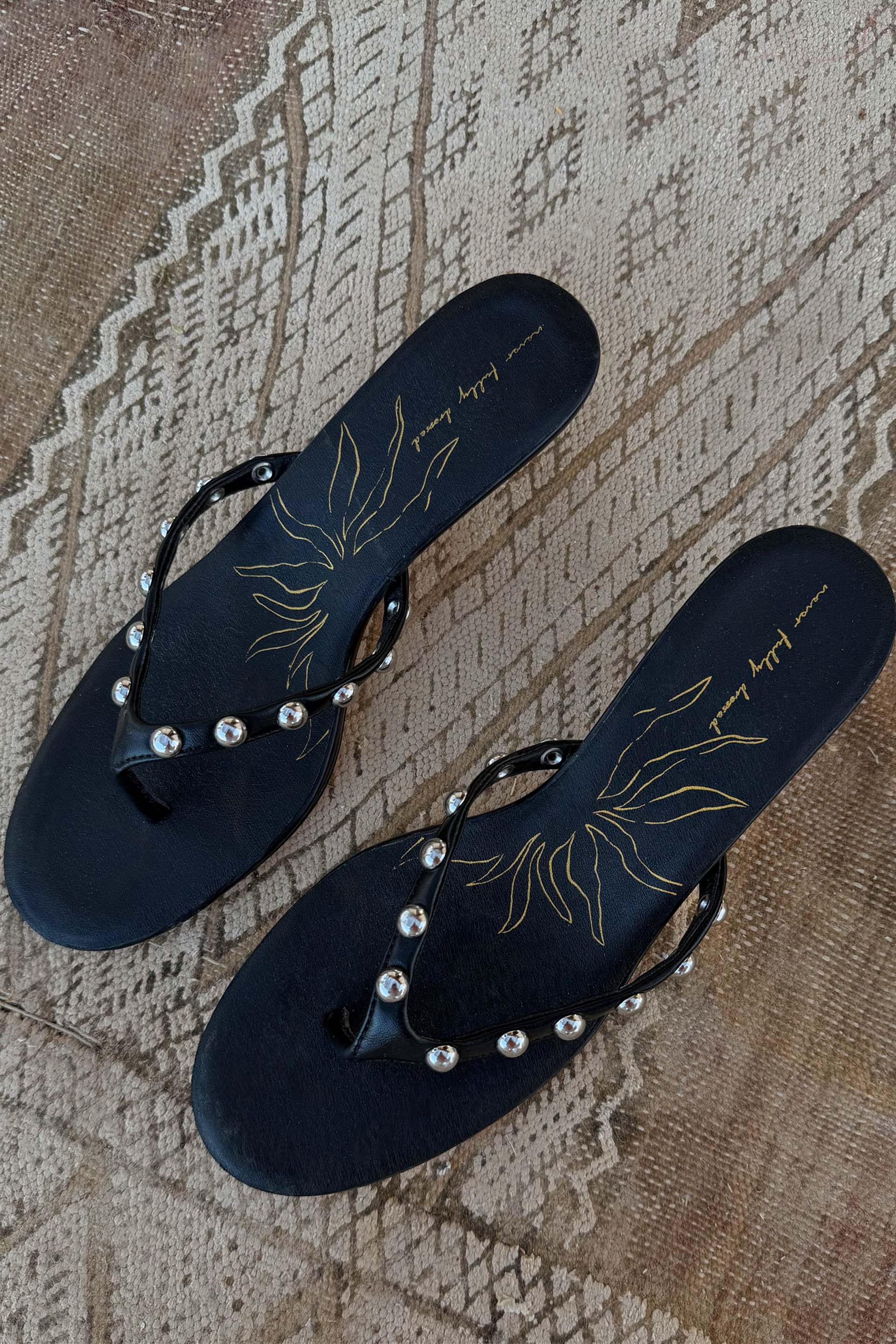 Black leather stud sandal mules with silver detailing and a gold sun motif, displayed top-down on a woven rug.