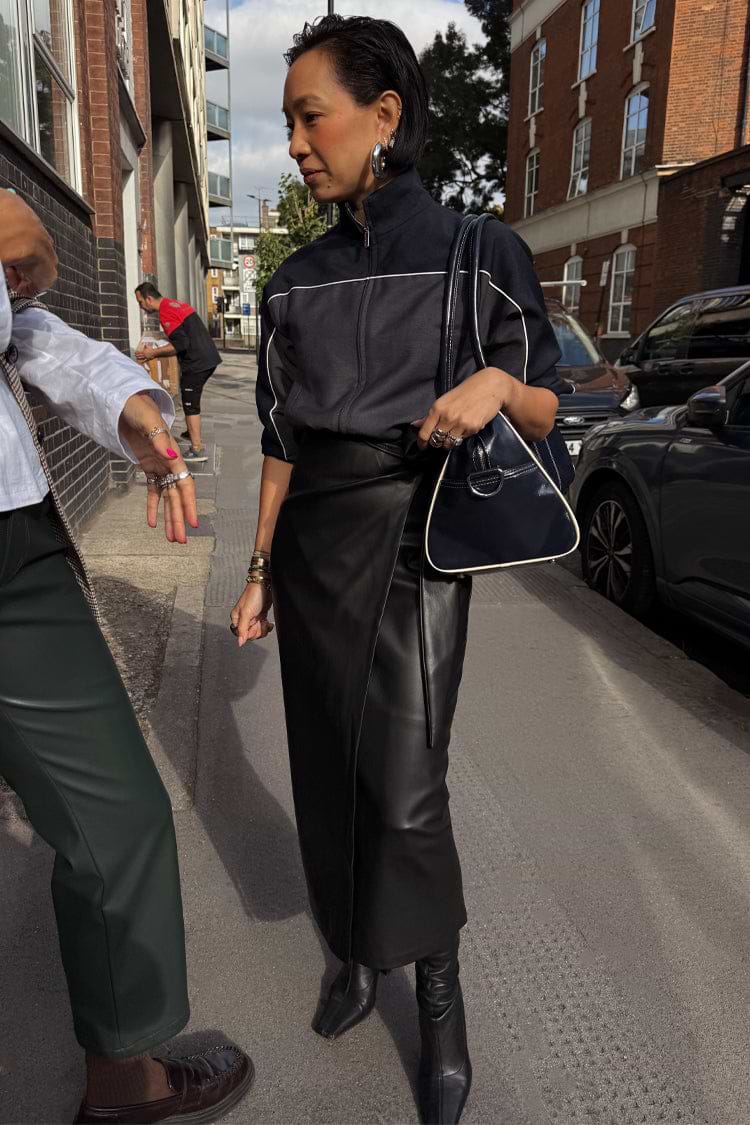 Black Vegan Leather Jaspre wrap skirt with a tulip shaped tie, paired with a zip-up jacket in a London setting.