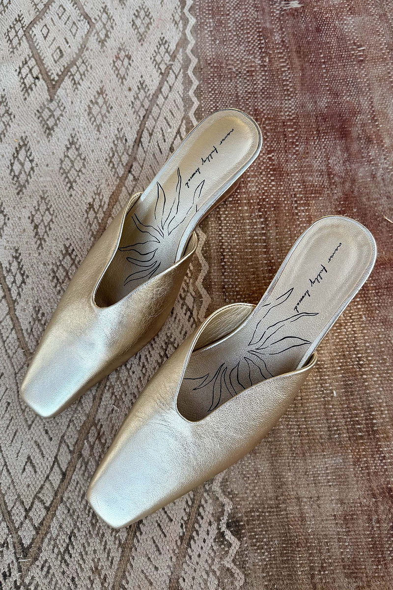 Gold metallic leather wedge mules with a pointed toe and open back, resting on a textured rug.