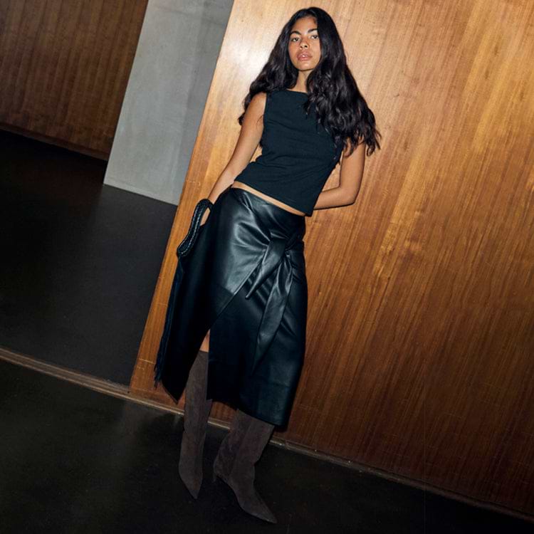 Black Vegan Leather Wrap Skirt featuring a tulip shaped tie, styled with a black top and tall brown boots.