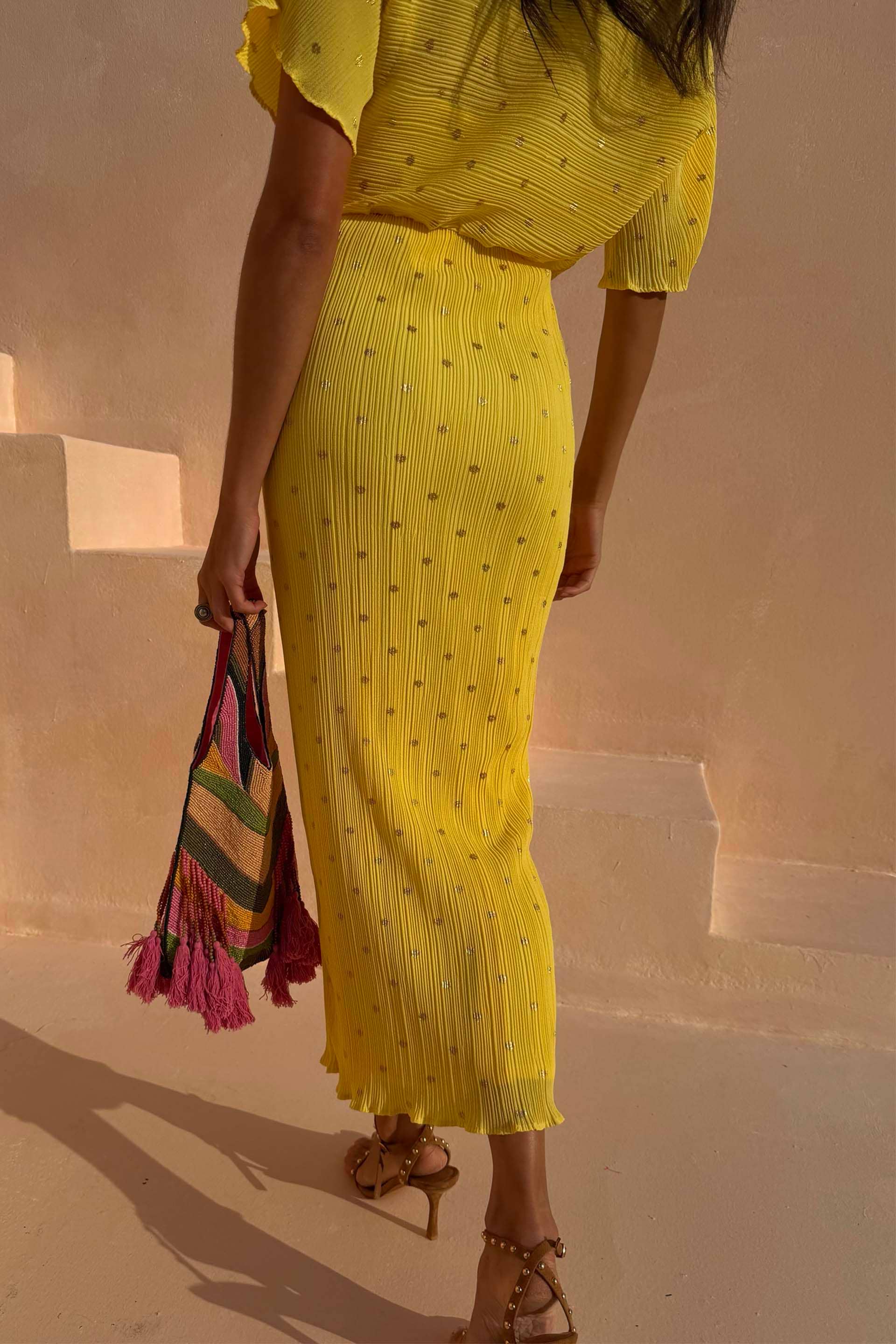 Sunshine Tilly Dress in yellow plissé with gold flecks and lettuce hem, held with a colourful beaded bag.