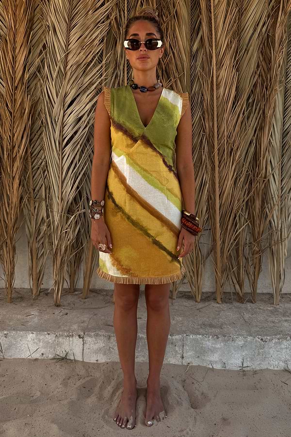 Green Ombre Deia Dress with yellow tones and raffia trim, styled for a vacation against palm leaves.