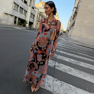 A woman stands on a city street crosswalk wearing a long-sleeve maxi dress with a colorful paisley and floral print in pink, orange, blue, and black. She is holding a small brown woven handbag and wearing pointed-toe heels.