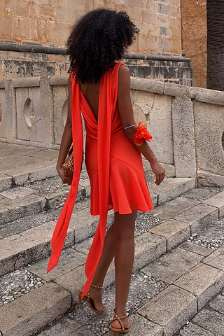 Tomato Mini Sophie Dress in red with a draped back and detachable scarf ties, shown on stone steps.