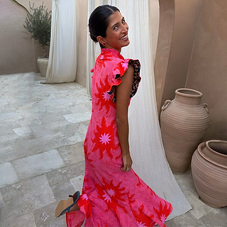 Pink Soleil Remi Dress with red sun motifs and leopard-lined pleated frills on a smiling woman in a courtyard.