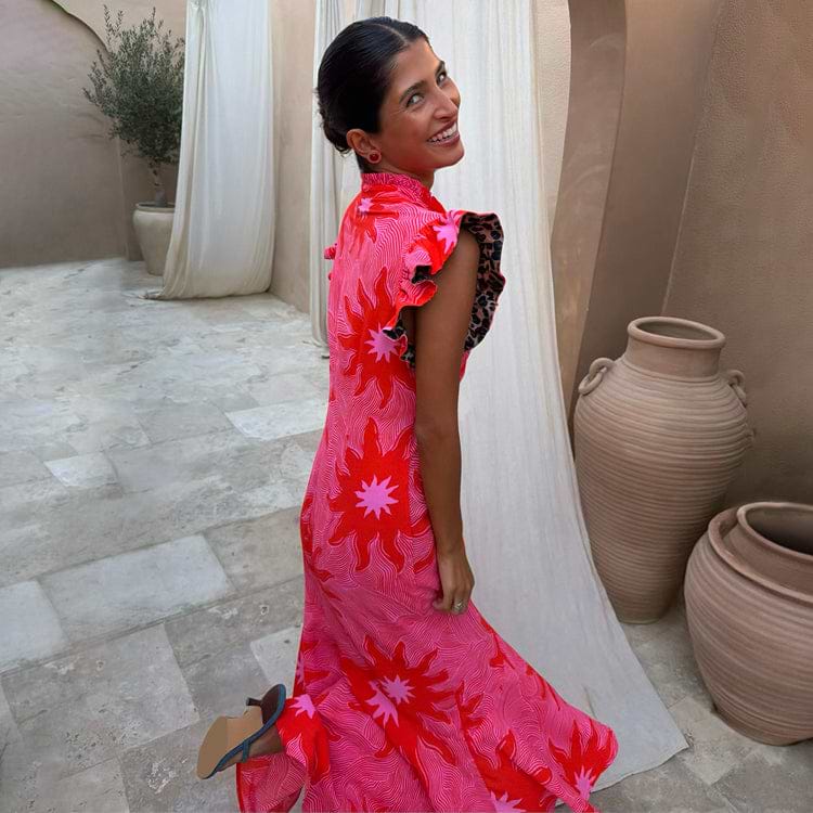 Pink Soleil Remi Dress with red sun motifs and leopard-lined pleated frills on a smiling woman in a courtyard.