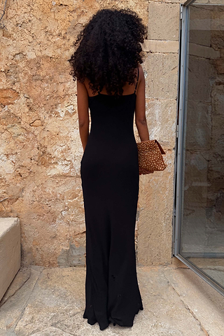 Sleek black Sapna maxi dress with adjustable straps and a fitted silhouette, shown from the back against a stone wall.