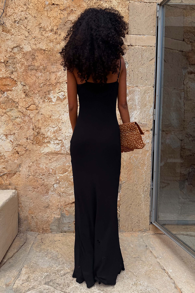 Sleek black Sapna maxi dress with adjustable straps and a fitted silhouette, shown from the back against a stone wall.