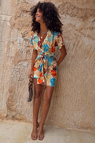 Shell Mini Wrap Dress featuring an abstract shell print, metallic detailing and flutter sleeves against a stone wall.