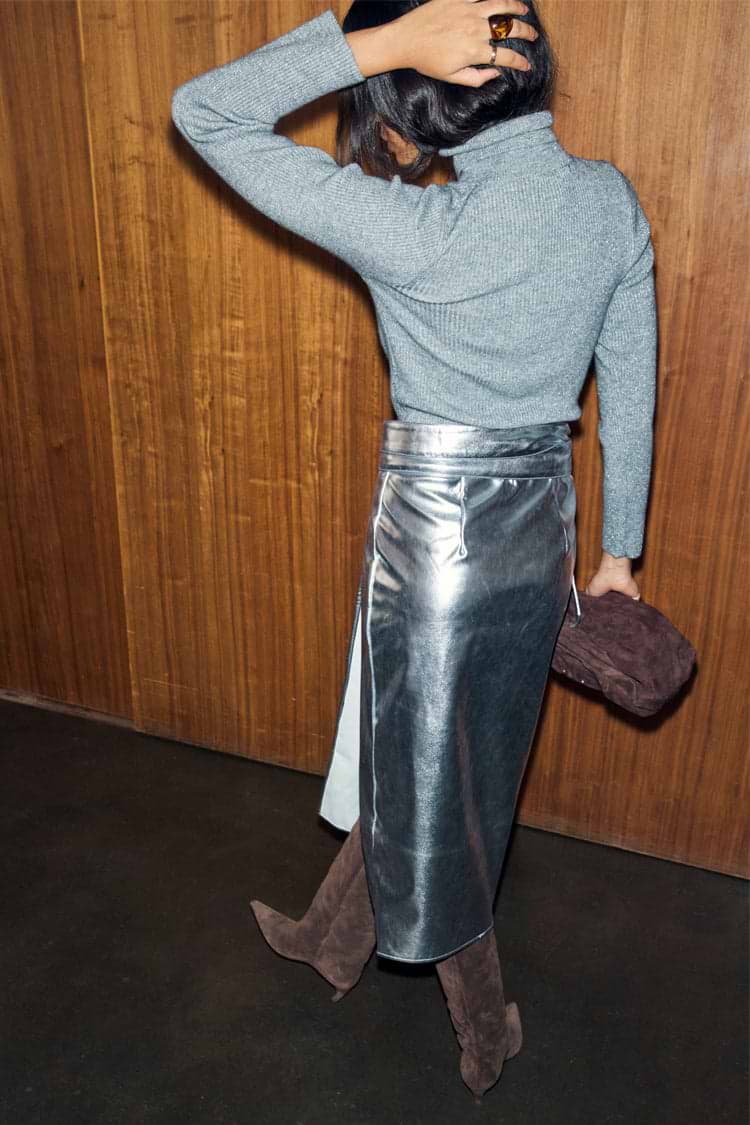 Silver Vegan Leather Jaspre Skirt with a grey knit and brown boots from behind against a wood wall.