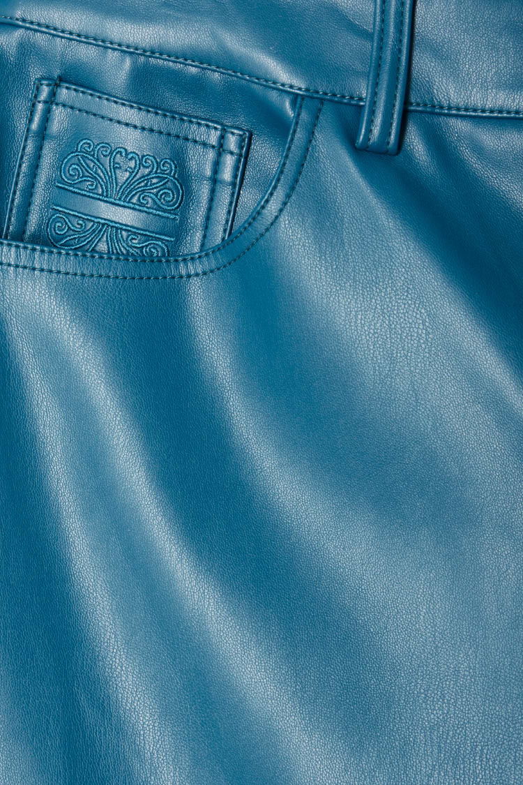 Close-up of teal vegan leather trousers featuring a detailed embroidered logo on the pocket and stitching.
