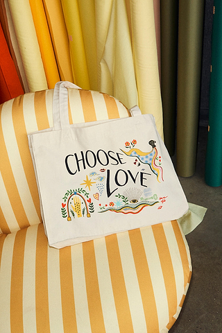 Choose Love Tote Bag in cream recycled cotton with colourful graphic prints, resting on a yellow striped armchair.