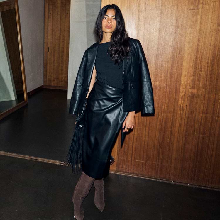 Black leather blazer draped over the shoulders, styled with a matching midi skirt and brown suede boots.