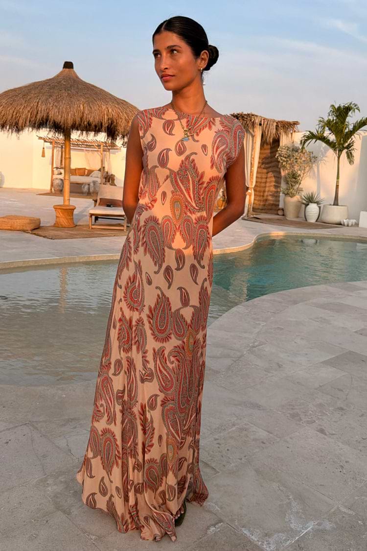 Peach Paisley Ava Dress, a bias-cut maxi with an all-over print, worn by a model in a poolside vacation setting.