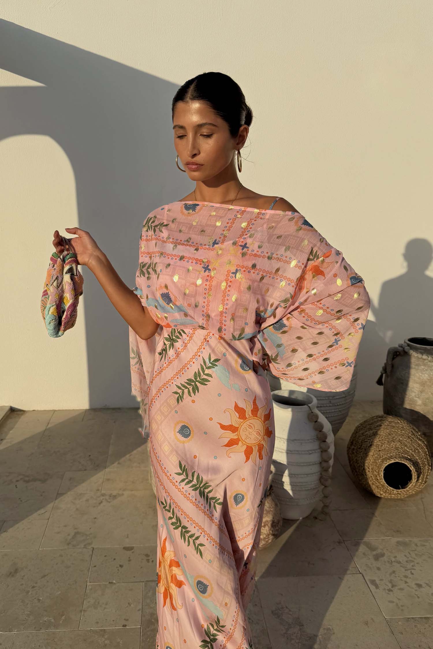 Pink Sun Anais Dress featuring a sheer draped top, gold sun motifs and a bias cut skirt on a sunny outdoor patio.