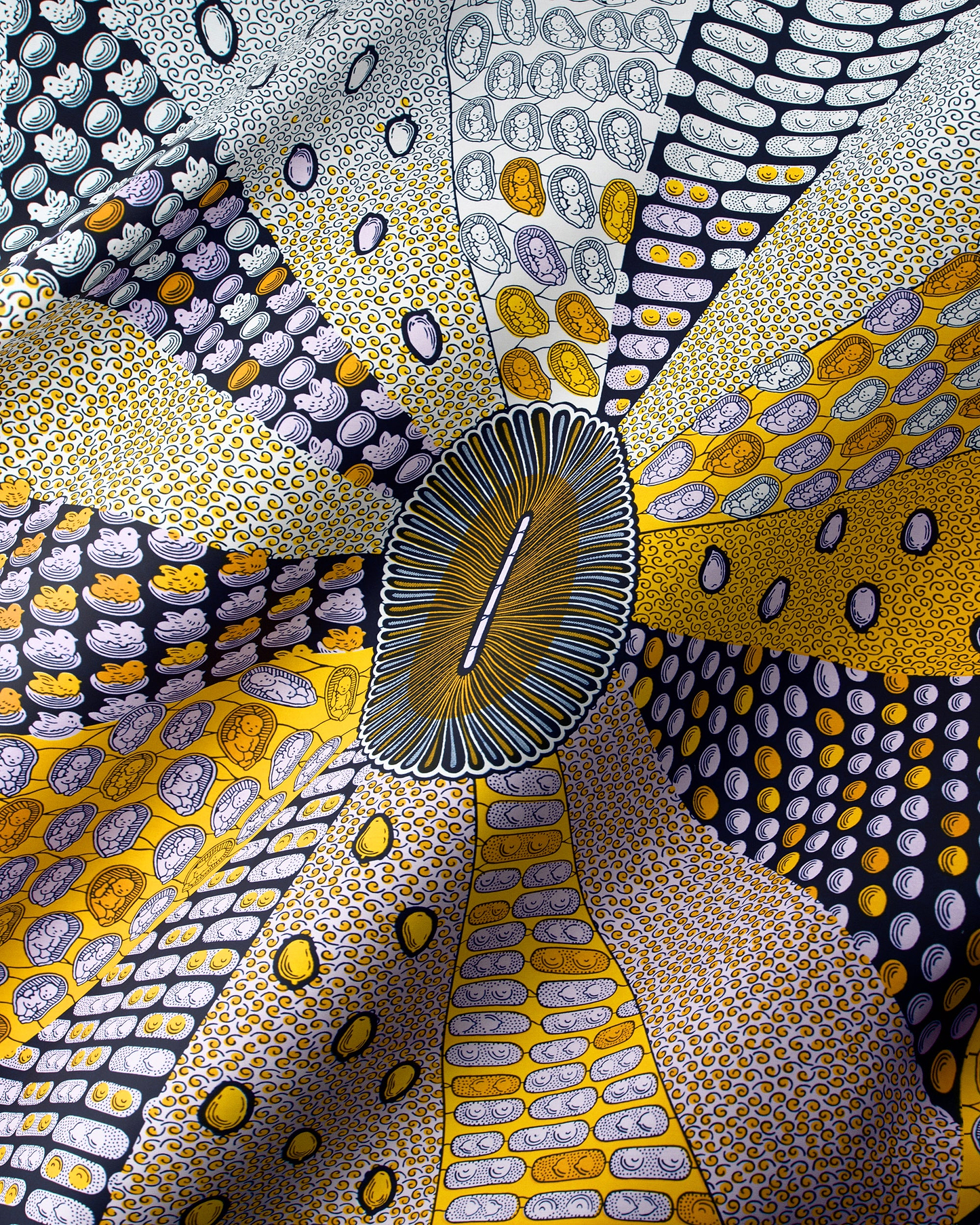 A close-up of a patterned fabric or textile with a radial, sunburst design. The material features multiple segments in yellow, gold, gray, black, and white, decorated with dense dot art and organic oval motifs, all radiating from a central oval patch.