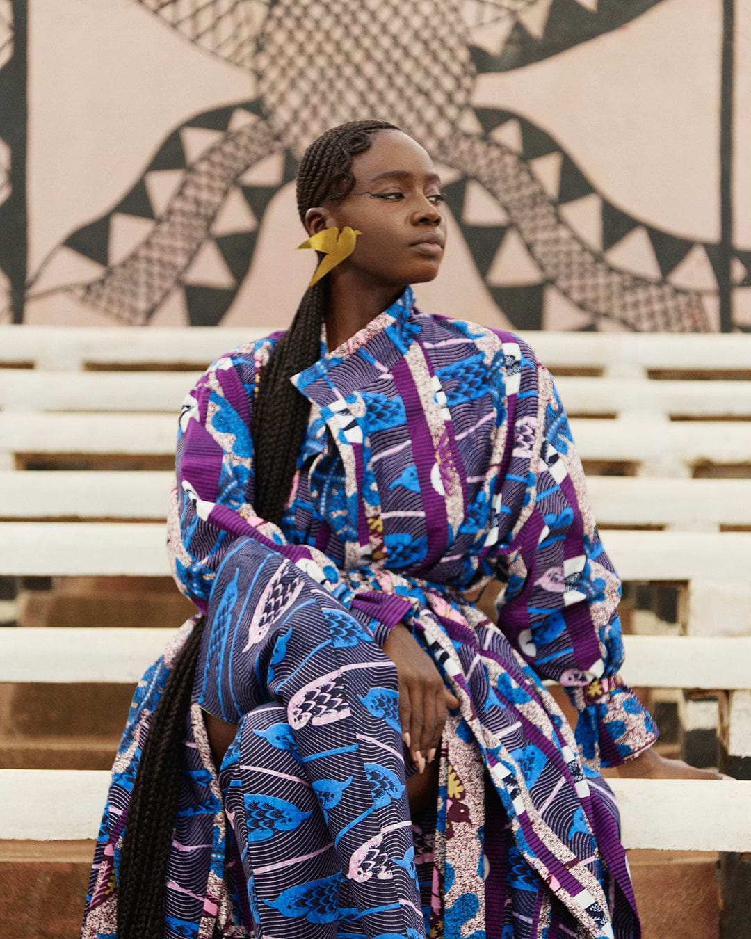 Sarah Diouf dressed in flowing Vlisco fabric with blue and purple bird patterns, seated on outdoor steps against a geometric mural backdrop