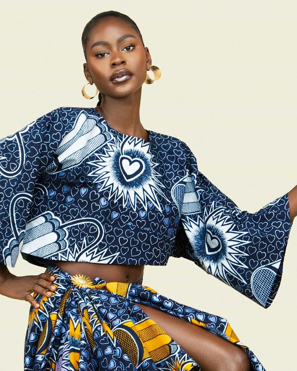A model wearing a matching two-piece African wax print set with a navy blue and yellow geometric pattern, set against a solid light beige background.
