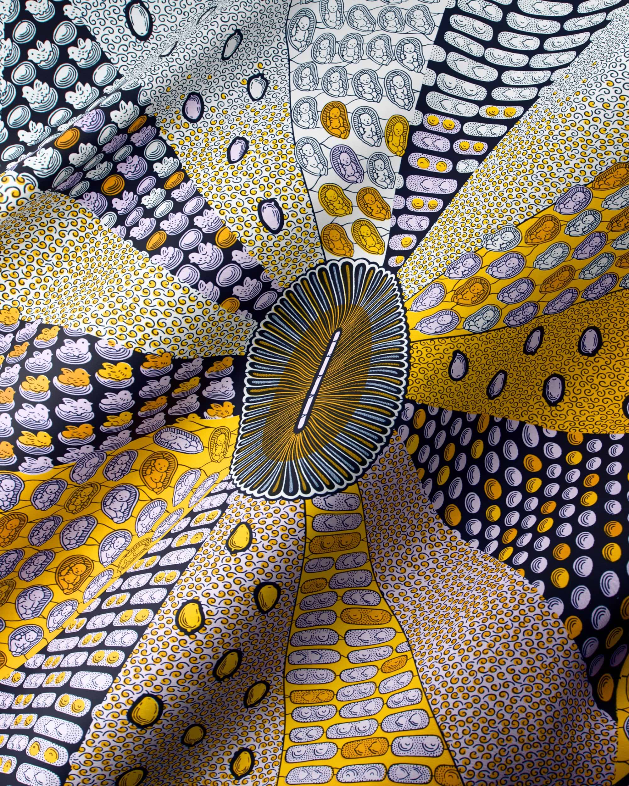 A close-up of a patterned fabric or textile with a radial, sunburst design. The material features multiple segments in yellow, gold, gray, black, and white, decorated with dense dot art and organic oval motifs, all radiating from a central oval patch.