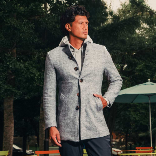 Man wearing a gray jacket and dark pants standing outdoors with tables and chairs in the background.