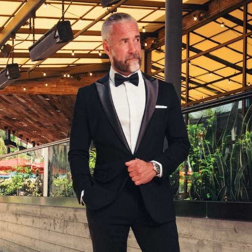 Man in a black tuxedo standing outdoors with plants and a building in the background