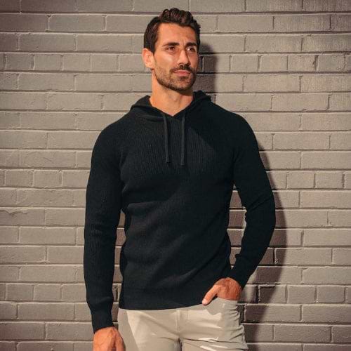 Man wearing a black hoodie and light gray pants against a brick wall.