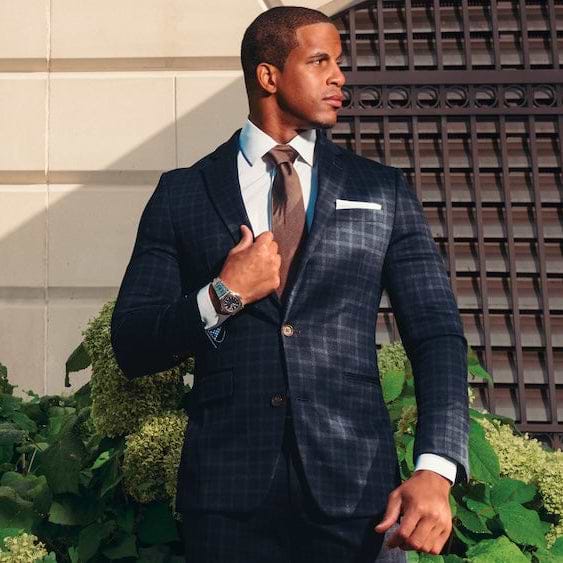Man in a suit standing outdoors with greenery and a building in the background