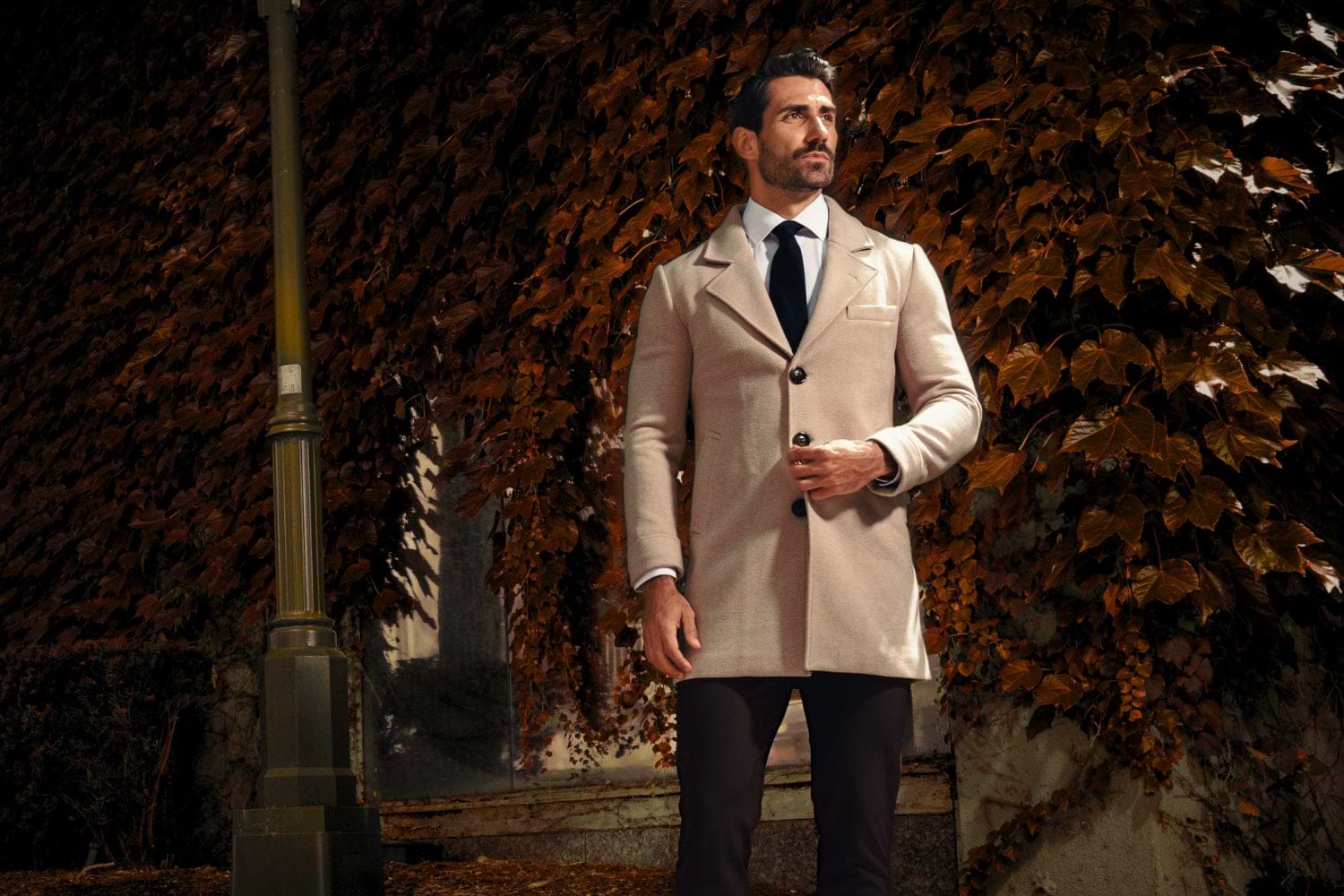 Man in a beige trench coat standing against a textured wall with autumn leaves.