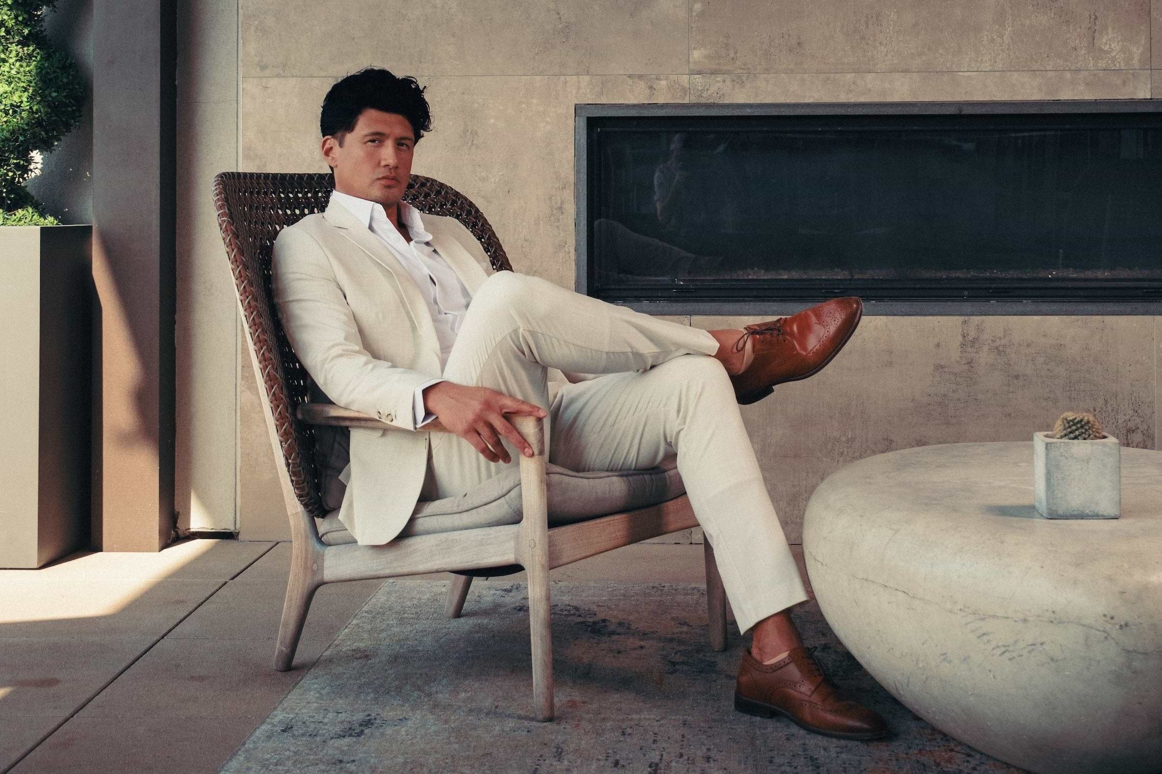 Man in a beige suit sitting in a modern outdoor setting with a fireplace.