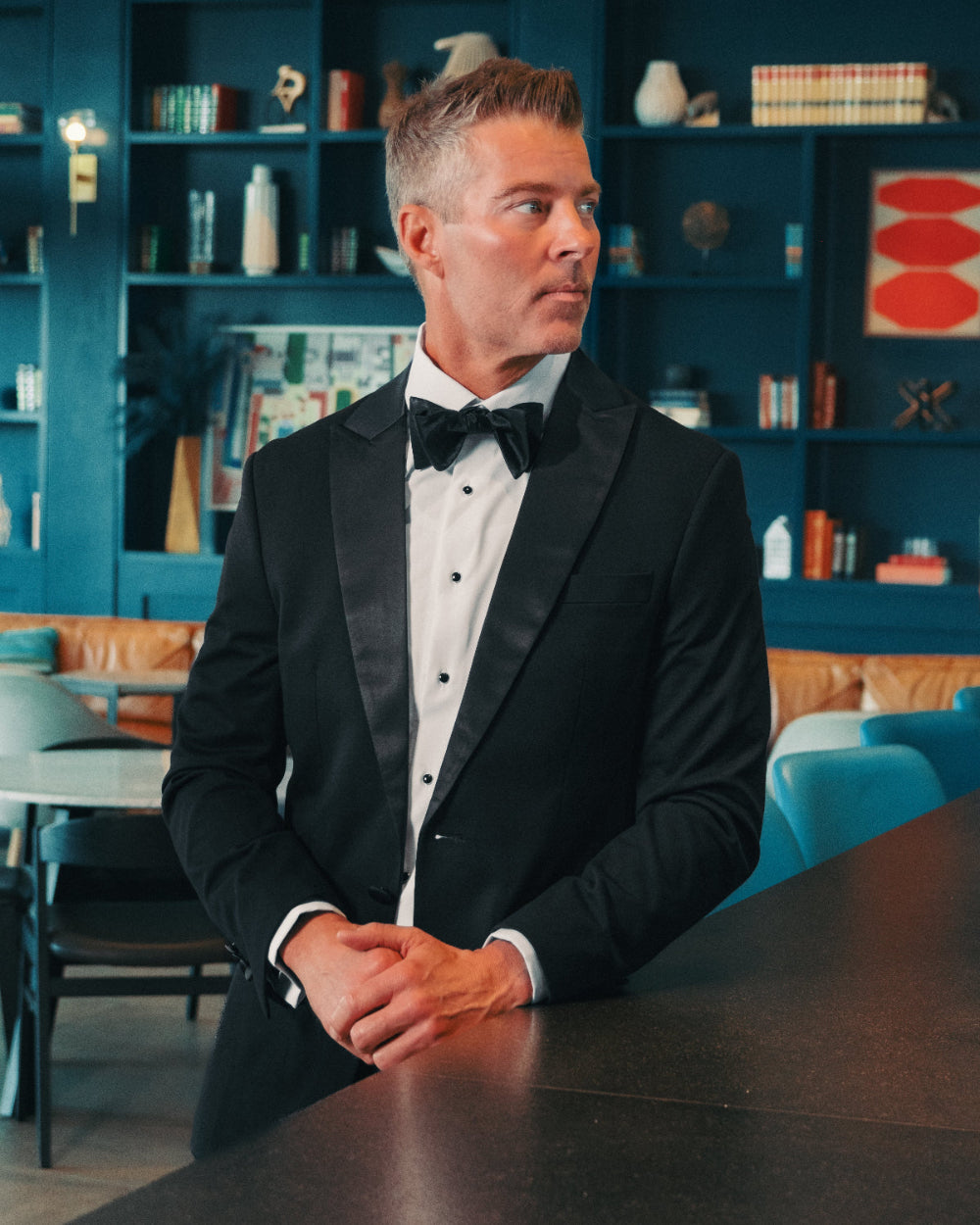 Man in a black tuxedo sitting at a table with a blue wall and shelves in the background