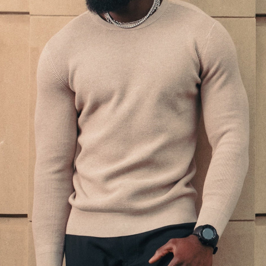 Man wearing a beige sweater and black pants standing against a modern building.