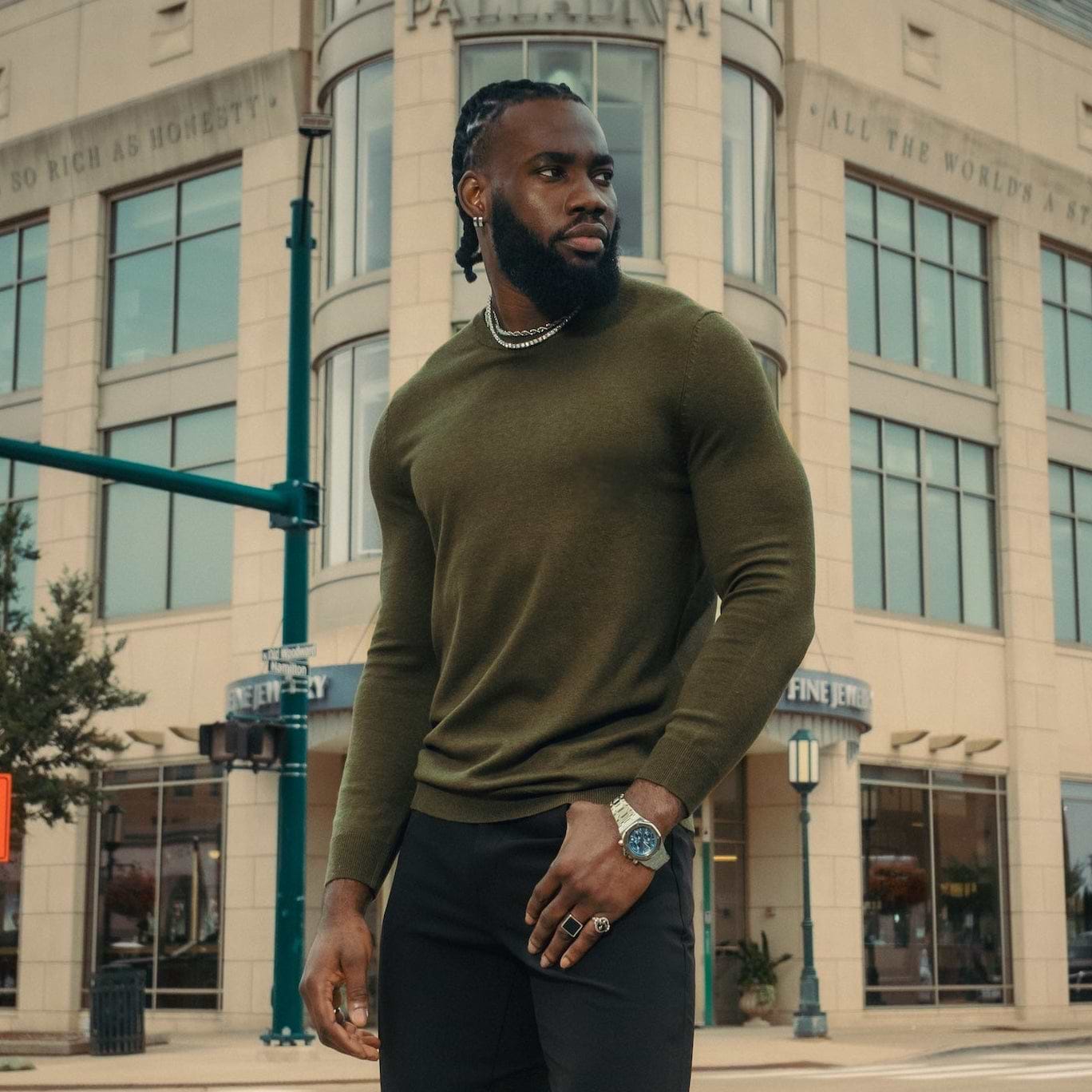 Man in green sweater and black pants standing on a city street corner.