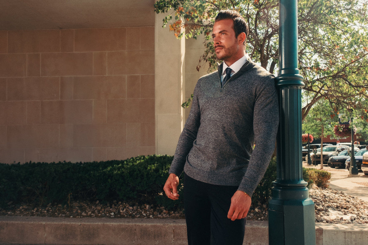 Man in a gray sweater and dark pants standing outdoors near a building.