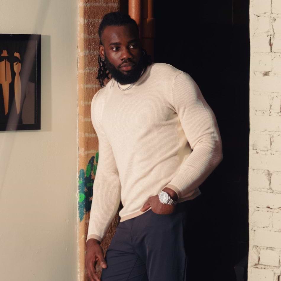 Man in a white sweater and navy pants standing on a staircase with a white wall and brick wall in the background.