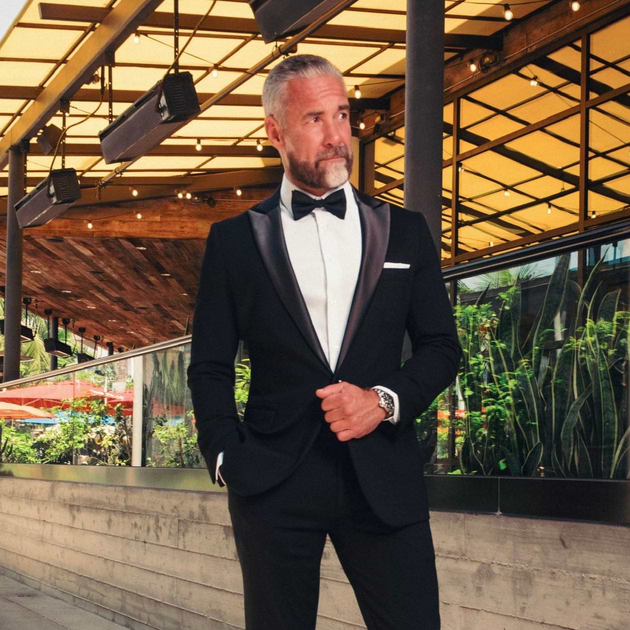 Man in a black tuxedo standing outdoors with plants and a building in the background