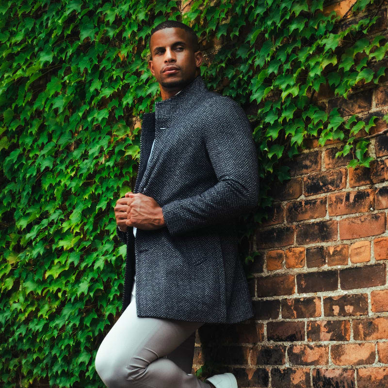 Man in gray sweater and pants leaning against a brick wall with greenery.