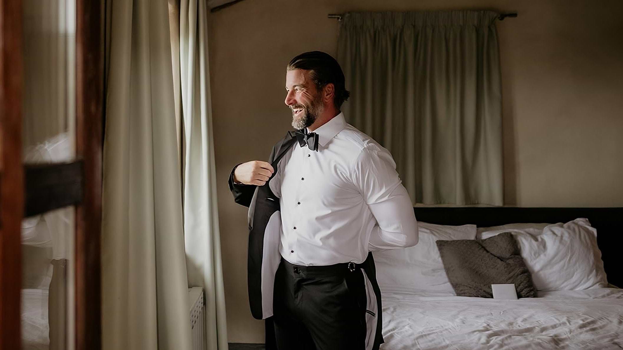 Man in formal attire standing in a bedroom with a bed and mirror.