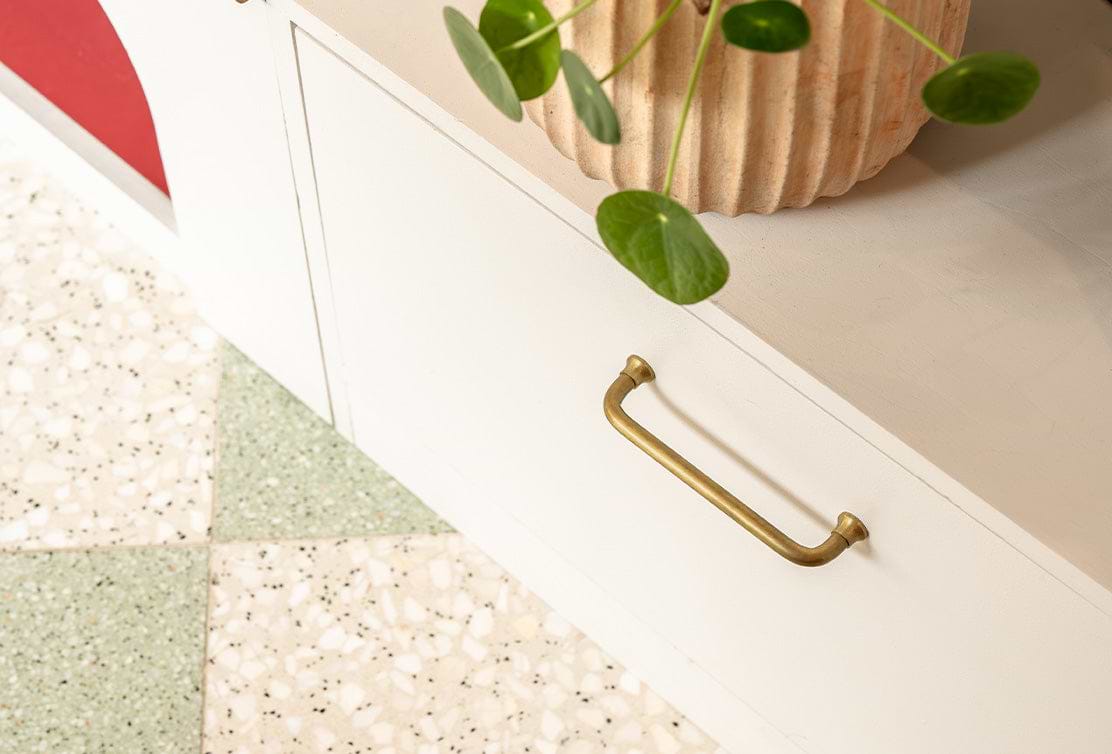A close-up of an antique brass 'Sycamore Furniture Handle' on a white cabinet with terrazzo flooring in the foreground.
