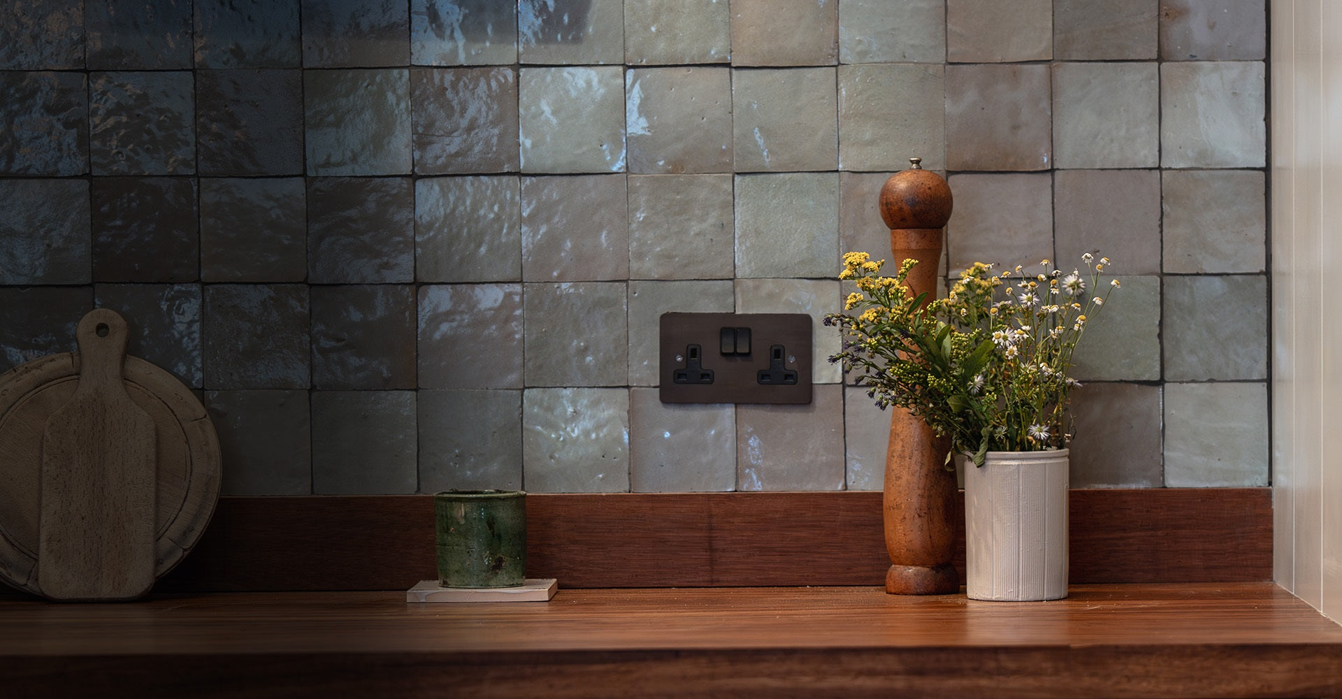 A rustic kitchen scene featuring a wood pepper mill, a vase of wildflowers, and a dark socket outlet against textured, muted tiles.