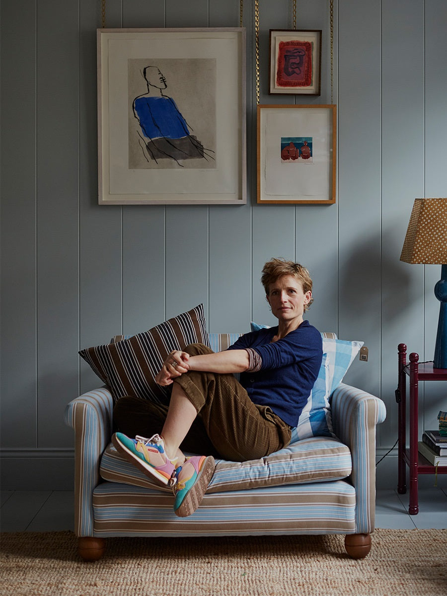 A person with short hair sits cross-legged on a striped sofa, surrounded by framed art and a patterned lamp in a cozy room.