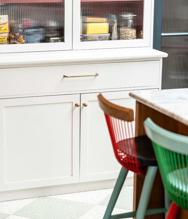 Modern kitchen cabinet with brass handles and colorful chairs in the foreground.