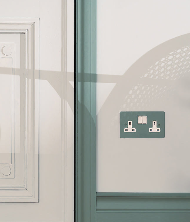 A close-up of a green wall socket against a white wall, with elegant molding and soft shadows highlighting the design.s29/05/2025