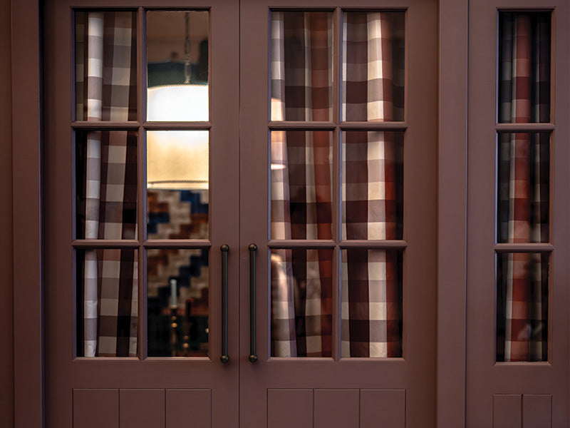 A pair of vintage-style doors with grid-pattern glass, framed by brown and cream checked curtains, revealing warm interior light.