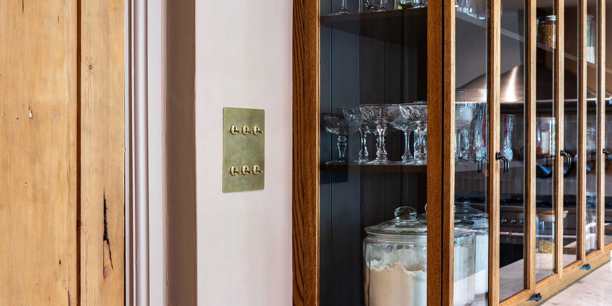 A wall-mounted brass switch plate with six toggles beside a wooden cabinet displaying crystal glassware and a glass jar.