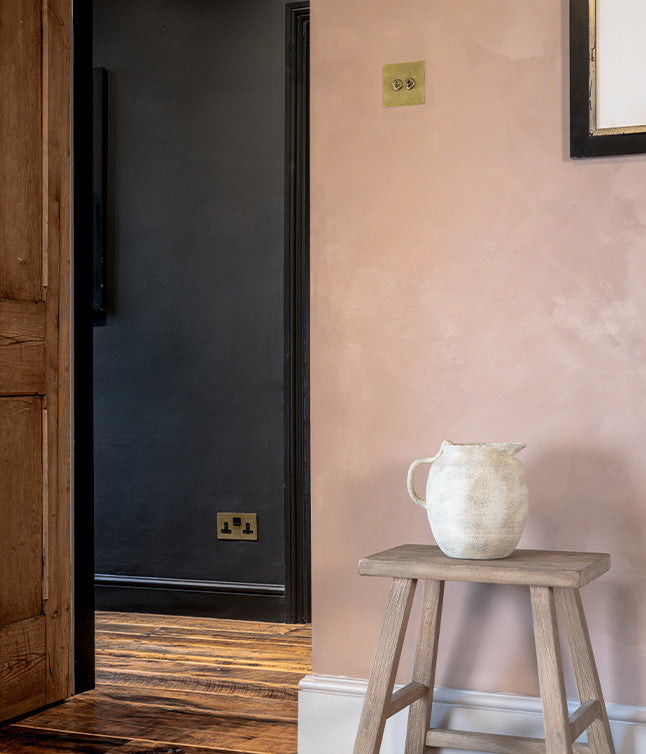 A minimalist interior featuring a wooden stool with a ceramic pitcher, soft pink wall, and a doorway to a dark room.