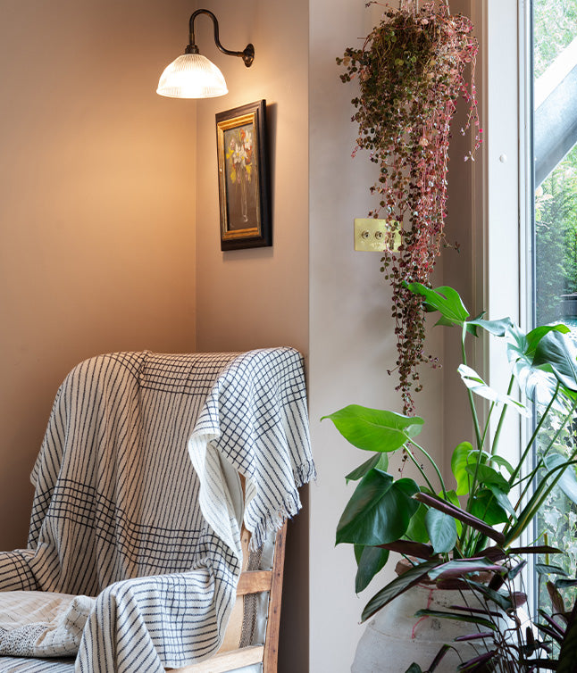 Cozy corner featuring a wooden chair draped with a checkered blanket, a wall lamp, a floral painting, and hanging plants by the window.