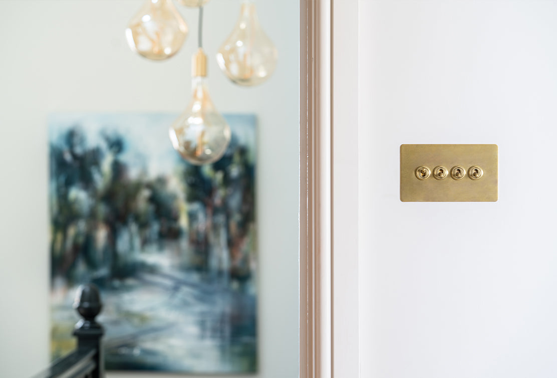 A close-up of a brass light switch panel on a white wall, with decorative pendant lights and a blurred landscape painting in the background.