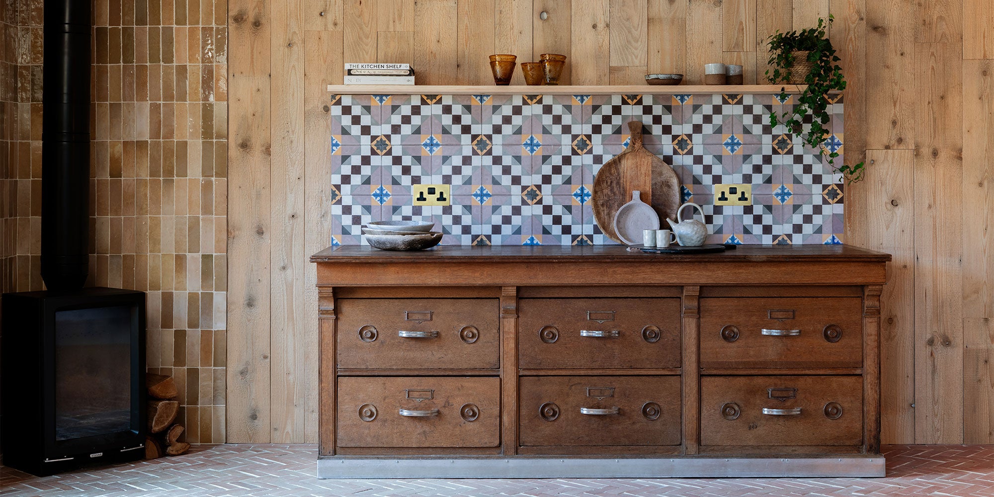 A wooden kitchen cabinet with a patterned tile backsplash, displaying various dishes, pots, and a decorative plant.