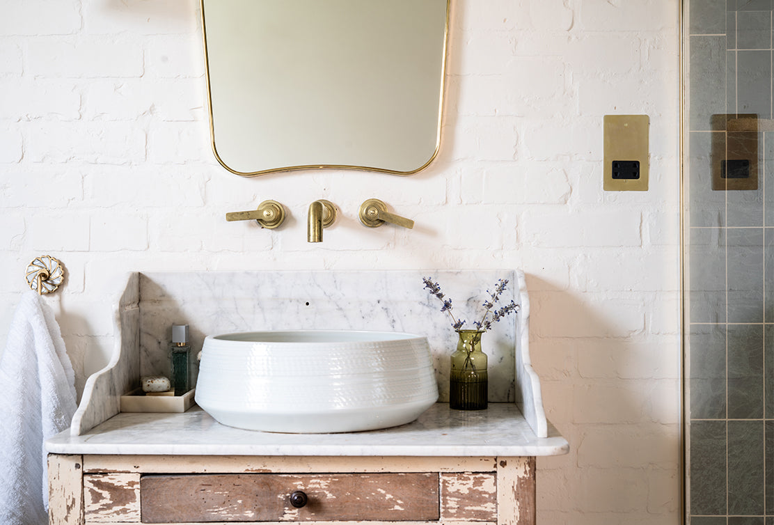 A chic bathroom sink with a round white basin atop a marble countertop, featuring gold faucets and a vintage vase with lavender.